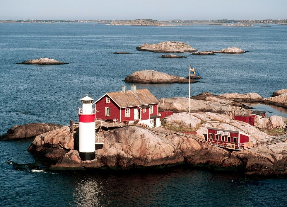 Aerial view of a small rocky island with a lighthouse, red house, and flag in the sea.