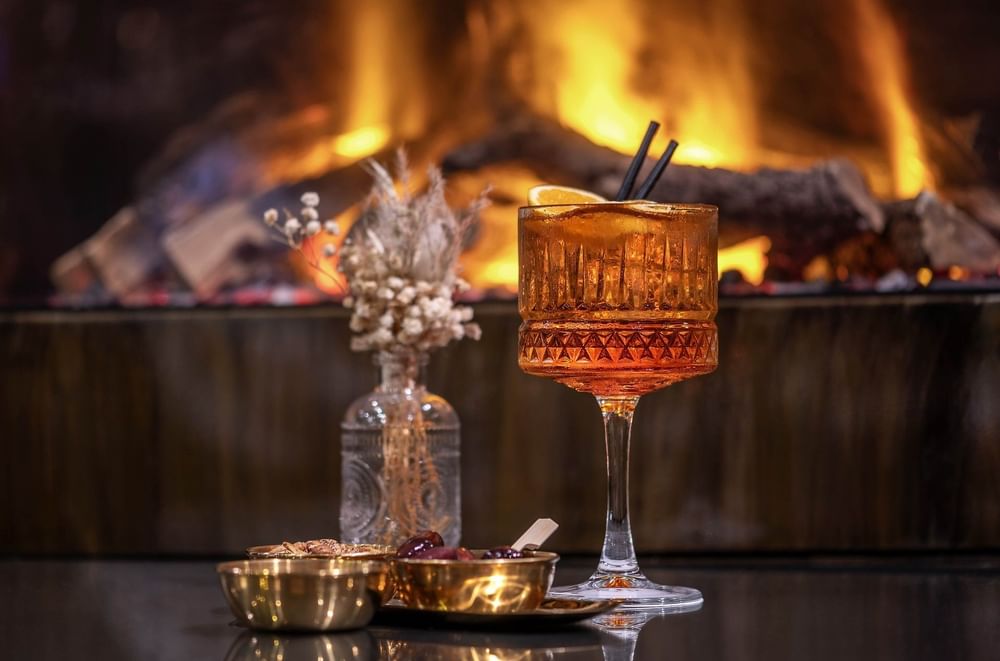 Amber cocktail in a textured glass set by gold bowls & dry floral vase under a glowing fireplace at Hotel Westminster Paris