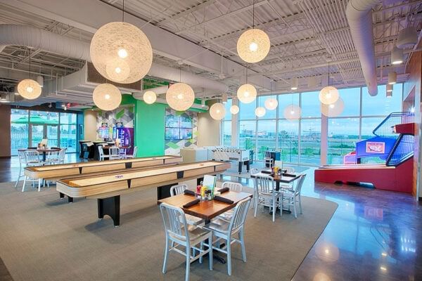 A modern game area with shuffleboard tables, dining tables, and chairs under large spherical lights.