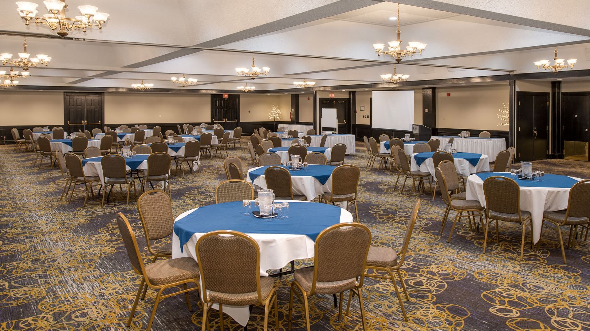 A banquet hall with round tables covered in white and blue tablecloths, each surrounded by multiple chairs.
