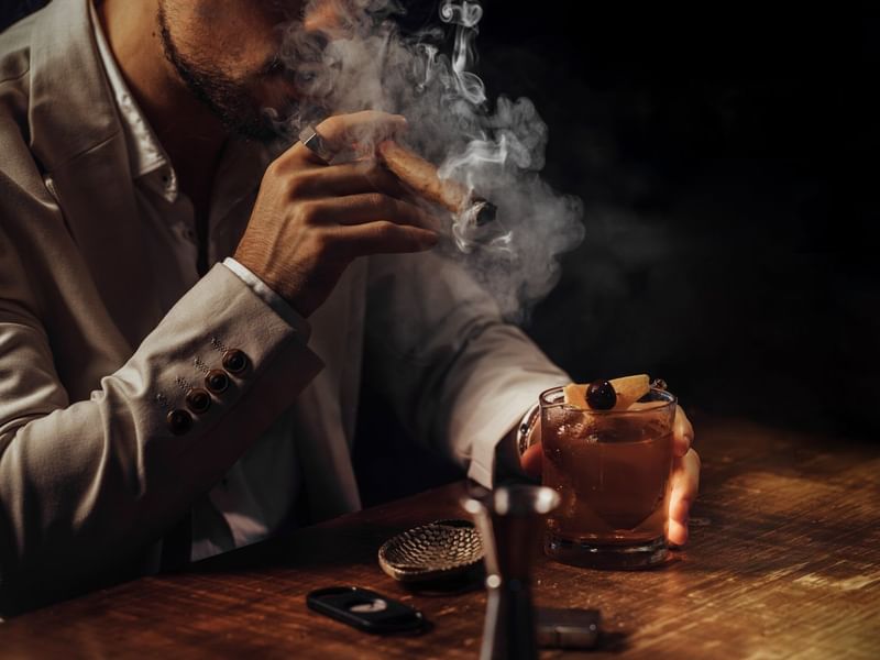Hombre sofisticado disfrutando un fino cigarro y cóctel artesanal en el lujoso lounge de Fiesta Americana Funeeq