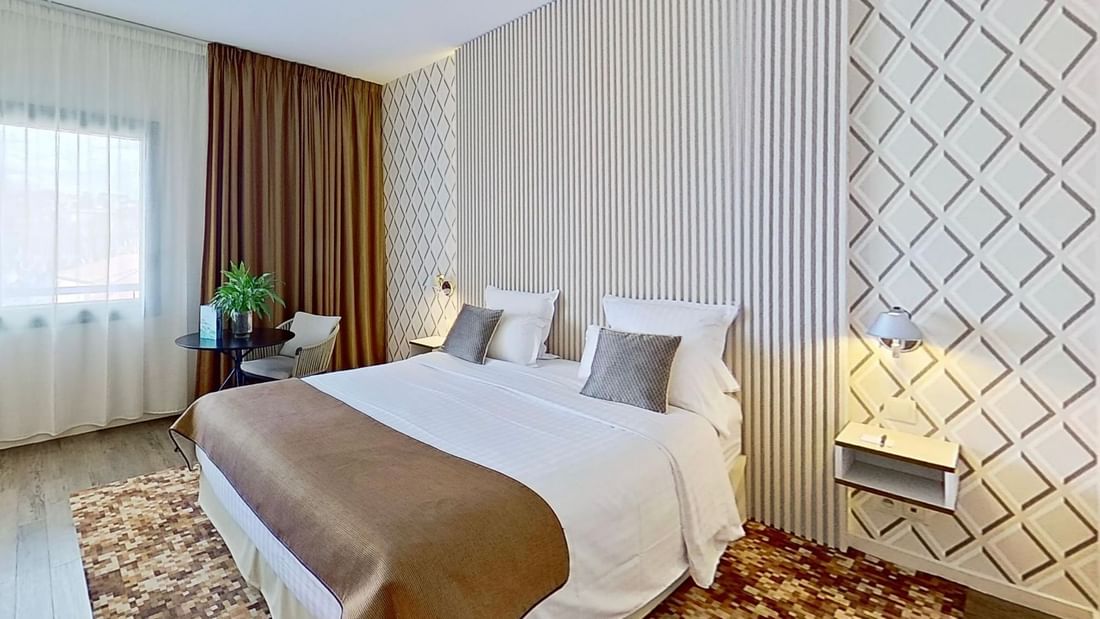 A hotel room featuring a large bed with white sheets, brown blanket, pillows, and a chair by the window.