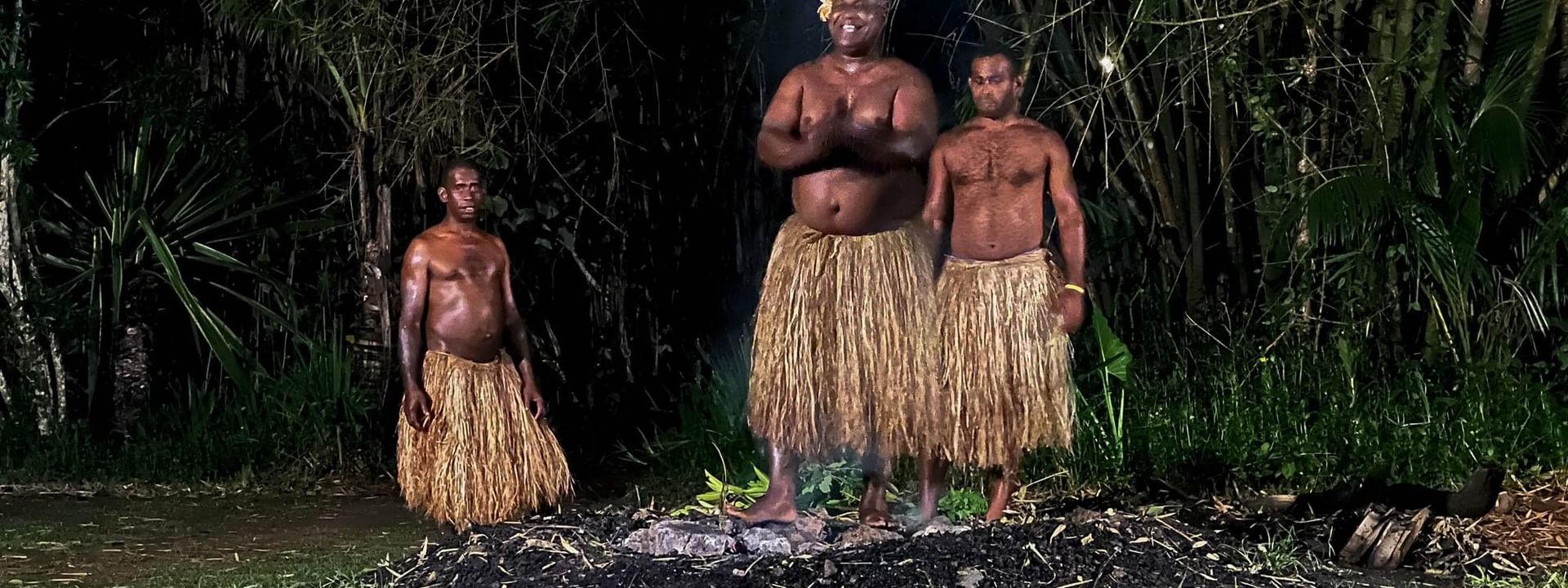 Three shirtless men in grass skirts stand near a fire at night in Korolevu at The Naviti Resort - Fiji