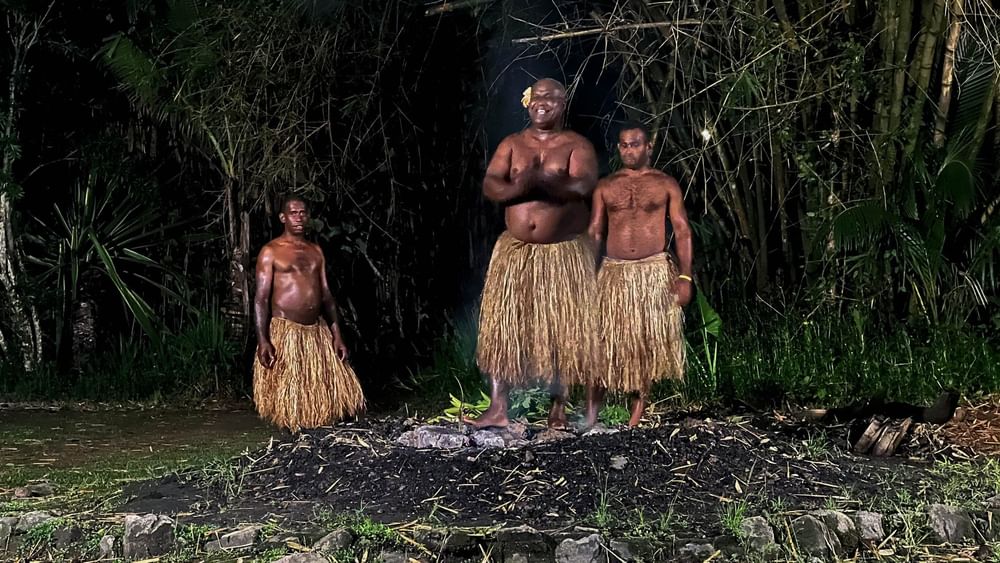 Three shirtless men in grass skirts stand near a fire at night in Korolevu at The Naviti Resort - Fiji.