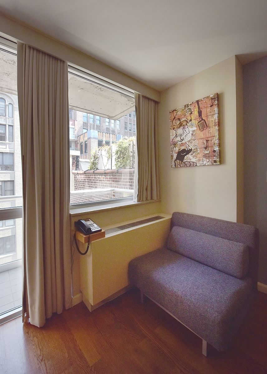 A room with a window, a gray sofa, a phone on a wooden shelf, and abstract artwork on the wall.