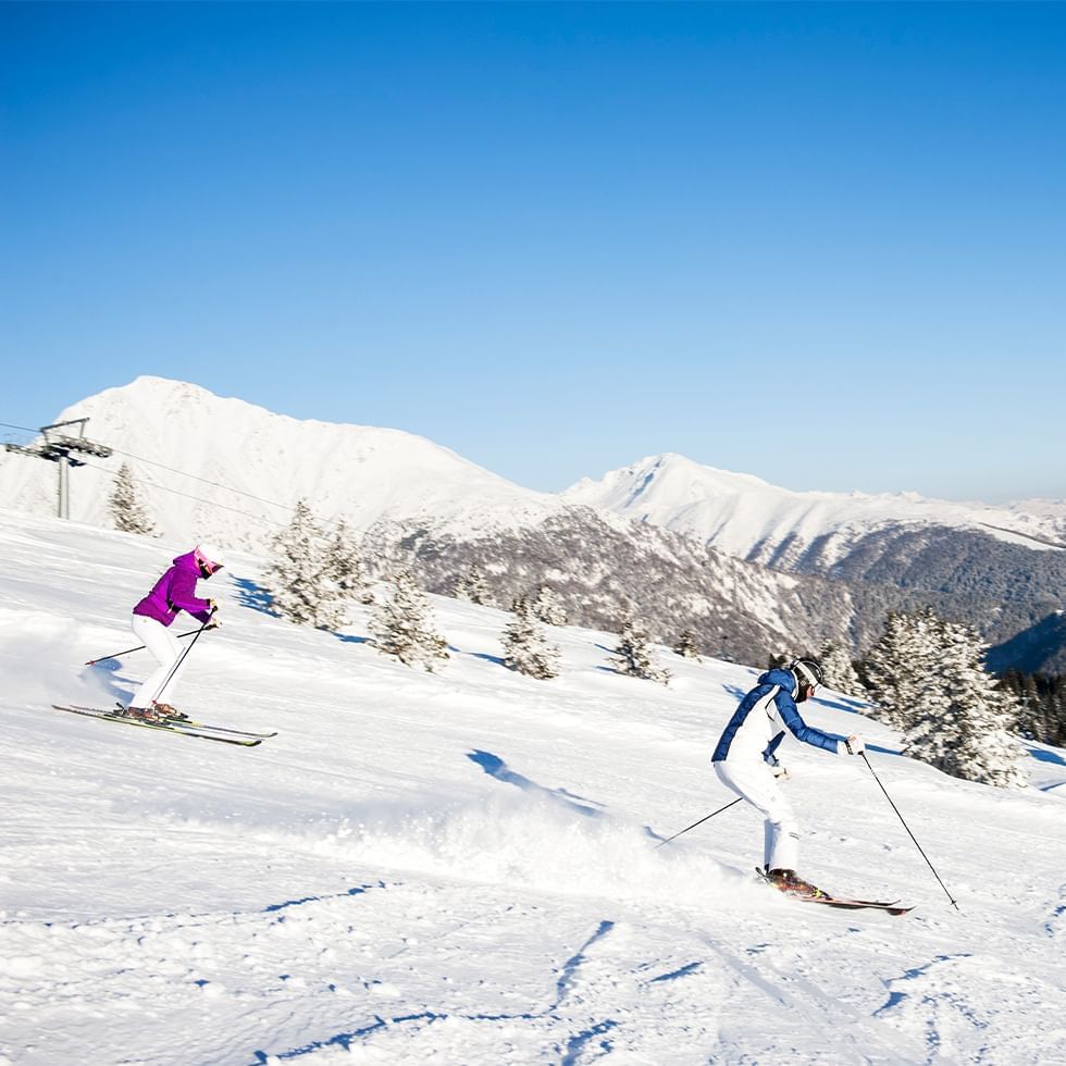 Zwei Skifahrer in bunten Skianzügen gleiten den schneebedeckten Berghang hinab.