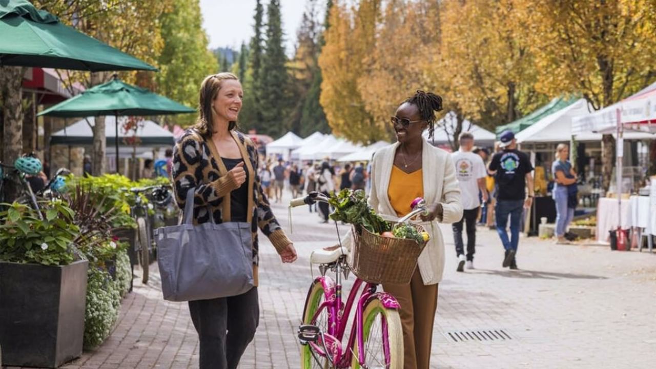 Two women stroll through an outdoor farmers market with a pink bicycle filled with fresh produce in Whistler