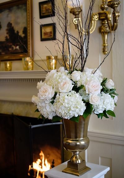 Bouquet of white roses and hydrangeas in a gold vase by a fireplace in the Willow Room at Spencer Hotel & Conference Centre
