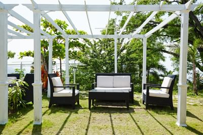 Black wicker sofa and armchairs under a white pergola, surrounded by greenery in Frank Room & Terrace at Hotel Montana Haiti