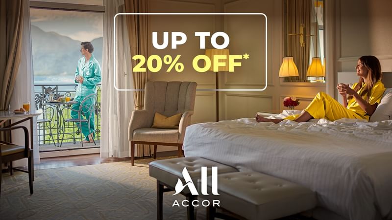 A hotel room with a woman relaxing on a bed and a man on a balcony.