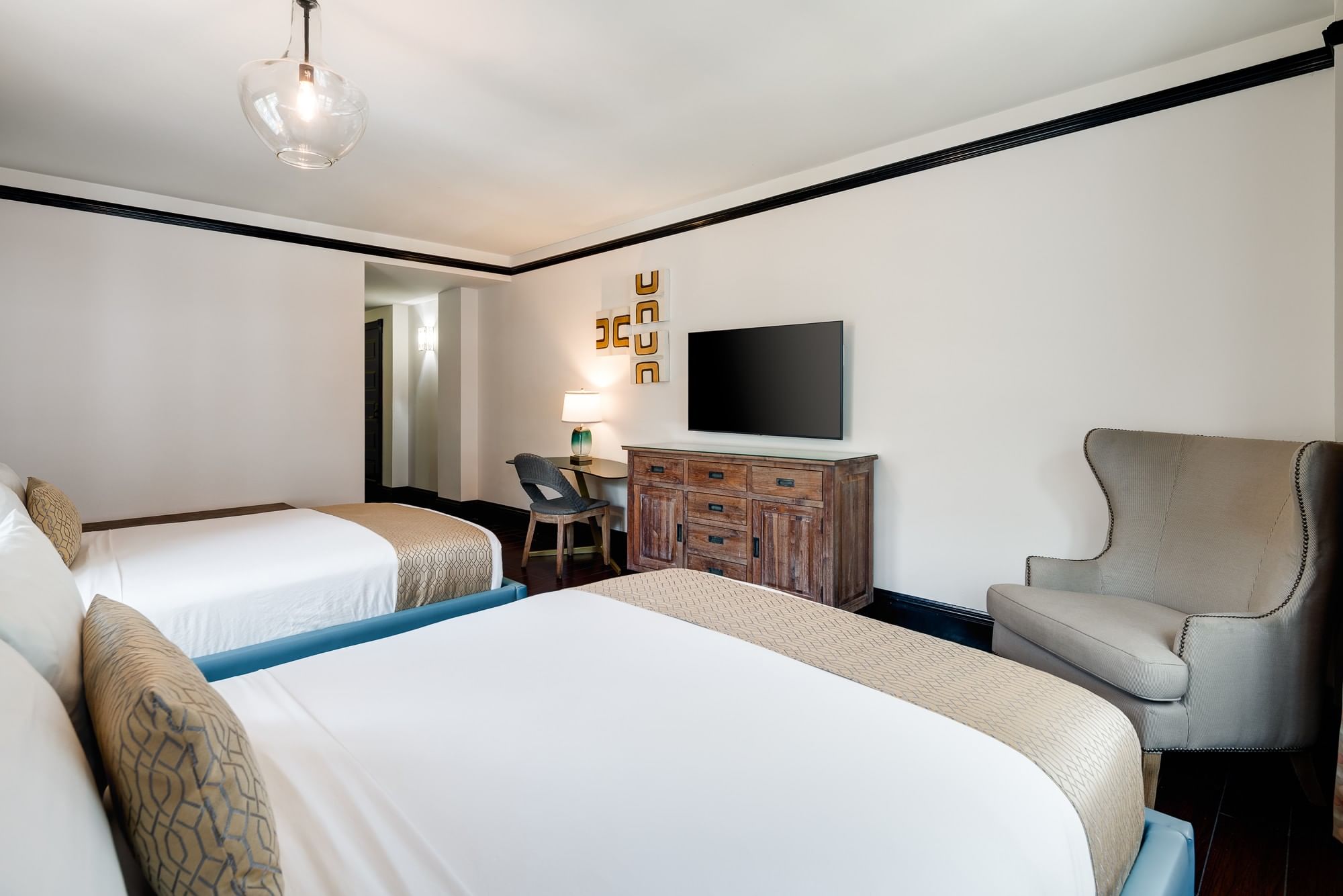 A hotel room with two beds, a wooden dresser with a TV on top, a desk with a chair, and a comfortable armchair.