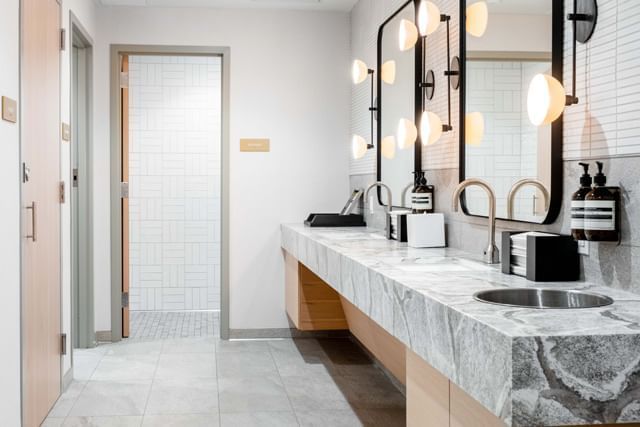 A modern bathroom with a double vanity, marble countertop, and lights over the mirror, with a shower in the background.