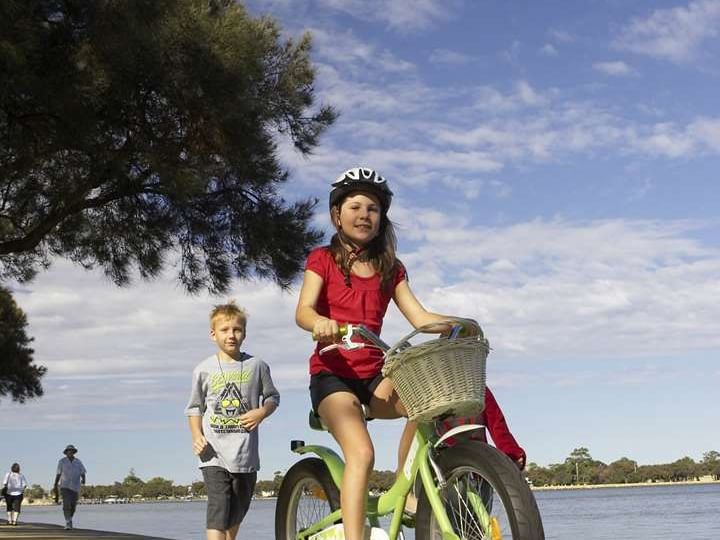 Two happy children cycling on a sunny day near the Sebel Mandurah boardwalk by the water