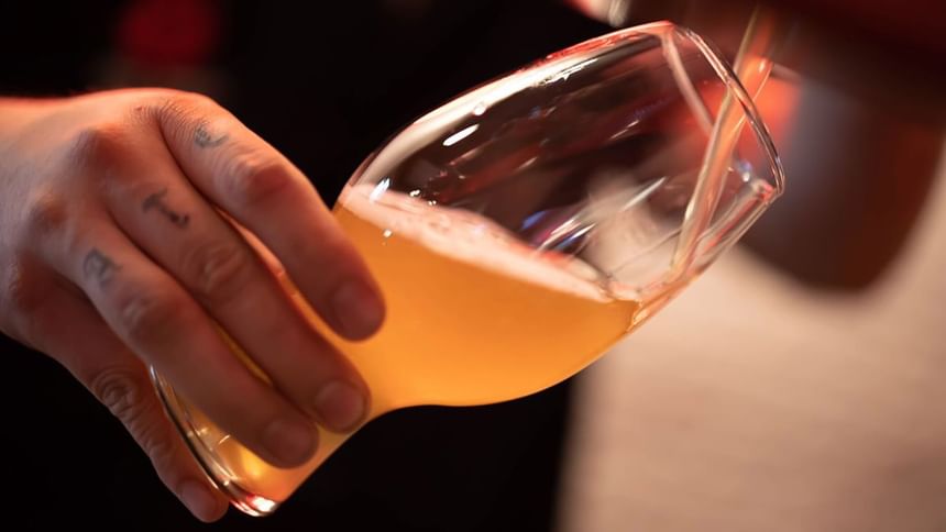 Close-up of a hand with finger tattoos pouring hazy beer into a unique glassware in Media Sangre at Araiza Calafia
