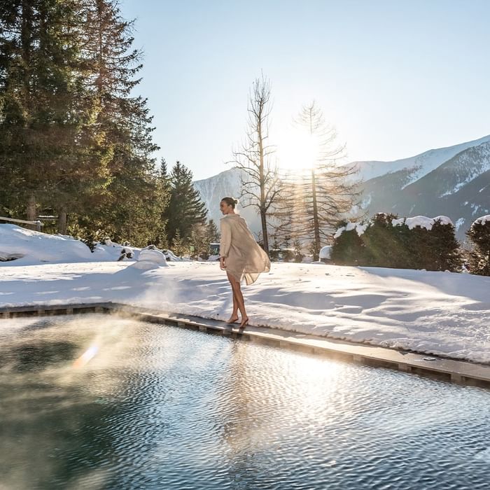 Frau in Bademantel neben einem Pool im Schnee mit Bergpanorama im Hintergrund.