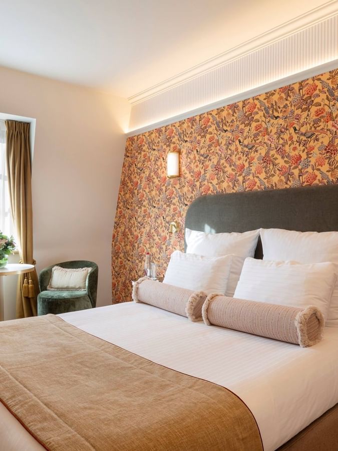 Moderne hotelkamer met kleurrijk bloemmotief behang, comfortabel bed en groene fauteuil bij het raam.