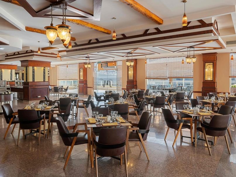 An empty restaurant with many tables and chairs, large windows, and decorative lighting fixtures on the ceiling.