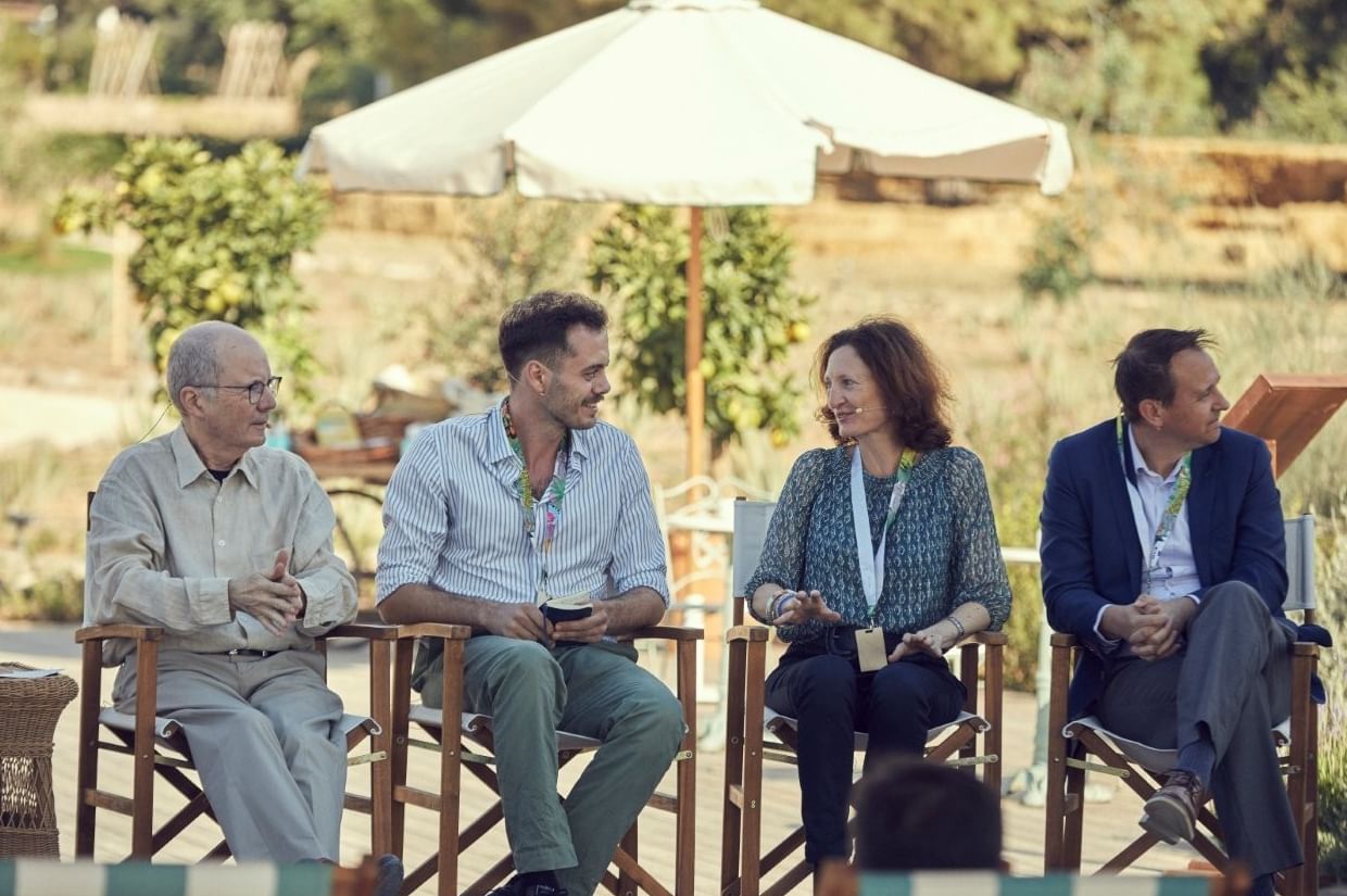 Four panelists seated outdoors under a large umbrella, chatting near citrus trees at Marbella Club resort