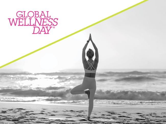 Woman practicing yoga in tree pose on beach at sunrise with Global Wellness Day logo and diagonal accent line