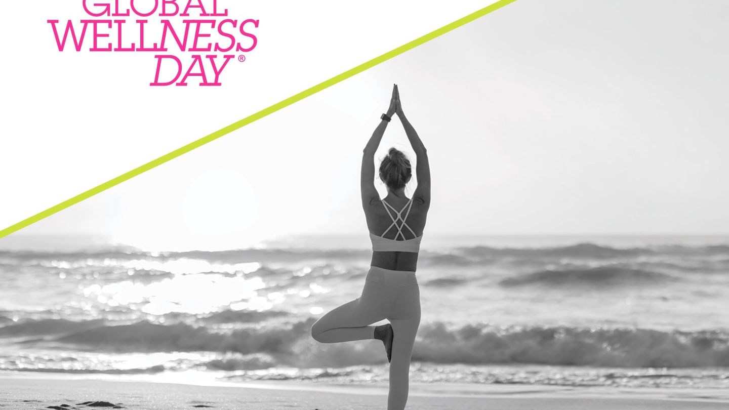 Woman practicing yoga in tree pose on beach at sunrise with Global Wellness Day logo and diagonal accent line