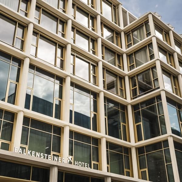 Facciata moderna dell'Hotel Falkensteiner con grandi finestre e logo in rilievo sotto un cielo azzurro.