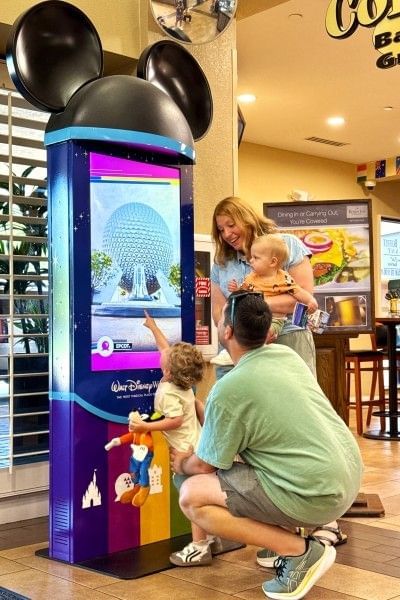 A mom, dad, and two young children stand in front of a Walt Disney World kiosk in the lobby at Rosen Inn Lake Buena Vista.