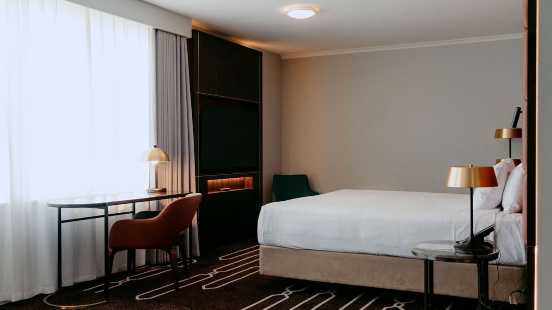 Modern hotel room with bed, desk, chair, TV, and window with curtains.