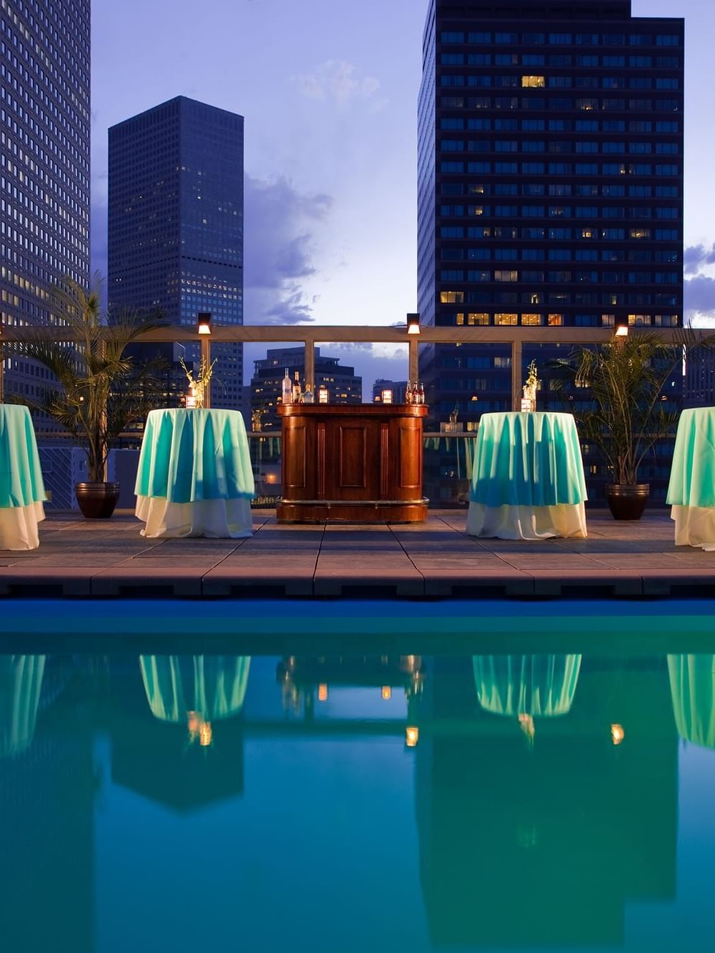 Small round tables with turquoise cloths by a rooftop pool under city buildings in Poolside at Warwick Denver