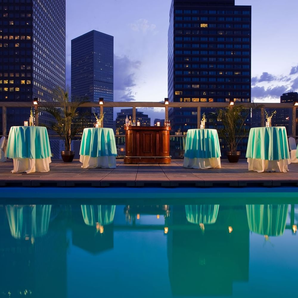 Small round tables with turquoise cloths by a rooftop pool under city buildings in Poolside at Warwick Denver