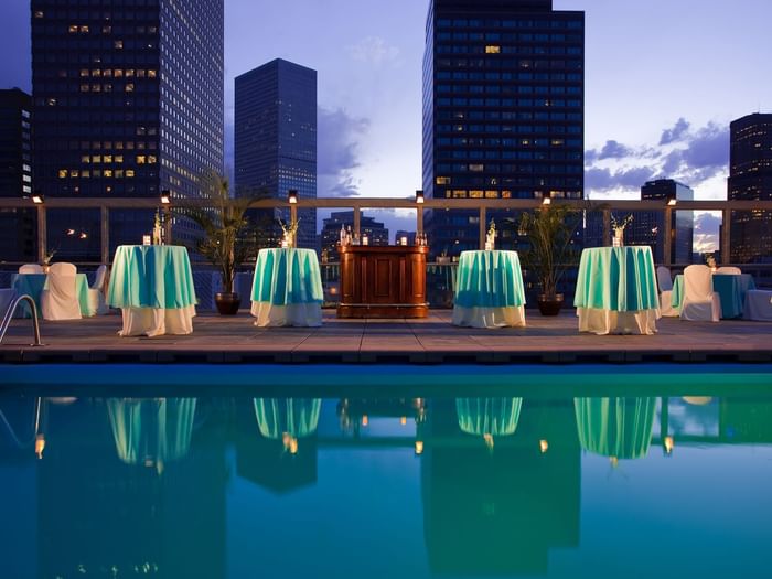 Small round tables with turquoise cloths by a rooftop pool under city buildings in Poolside at Warwick Denver