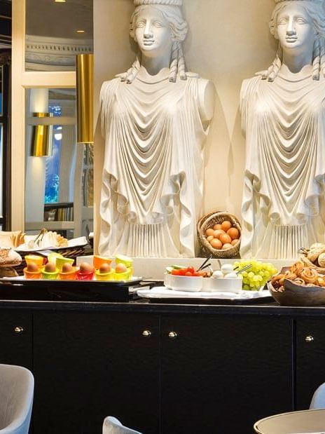Close-up of fresh bread and fruit with Greek Caryatid statues at the breakfast buffet in Hotel Barsey by Warwick