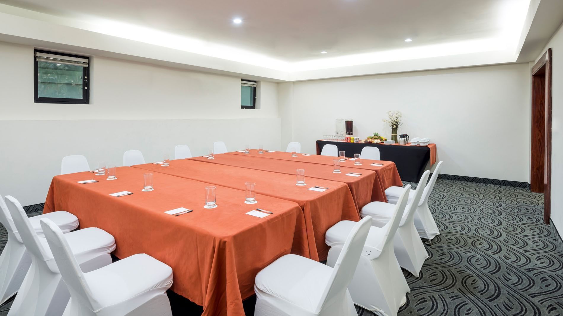 White chairs by a long orange table under recessed ceiling lights near a buffet table in the Budapest at Real Inn Mexicali