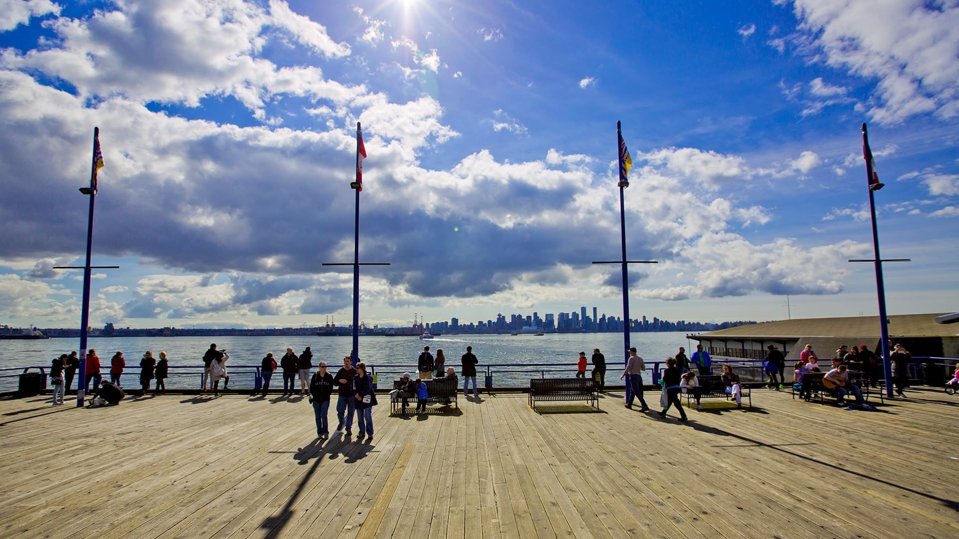 Wooden pier filled with people enjoying the view near the Coast Lonsdale Quay Hotel, with a city skyline in the background