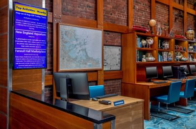 Computer workstations and concierge desk positioned by shelves with displays against a brick wall at Harborside Inn Boston