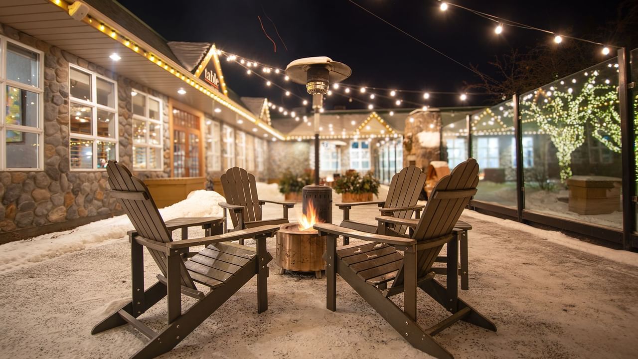 Three wooden chairs around a fire pit at night with string lights hanging above.