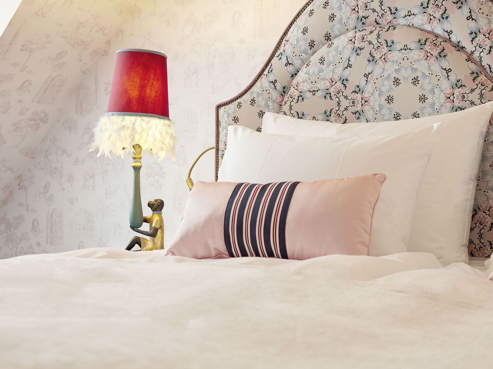 Bedroom with floral headboard, white bedding, pink striped pillow, and a red lamp with feather shade.