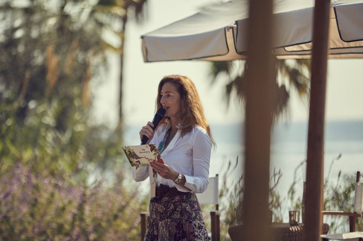 Woman holding a program and speaking into a microphone with the ocean blurred in the background near Marbella Club