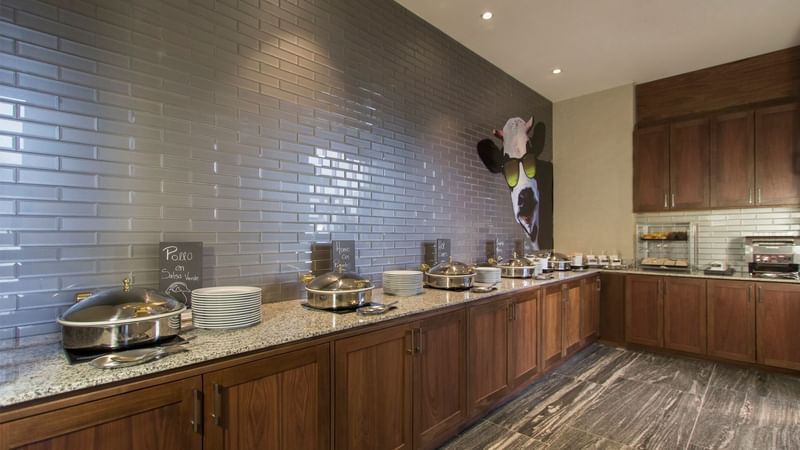 Buffet setup with silver trays and dishes, against a tiled wall at Fiesta Inn Suites Aeropuerto Del Bajío
