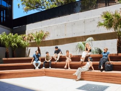 A group of people are sitting on wooden benches in a courtyard surrounded by palm trees.