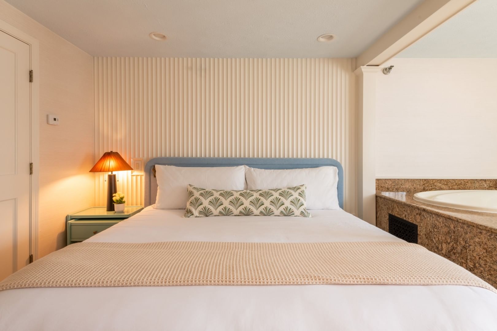 A neatly made bed with white linens and a decorative pillow, a nightstand with a lamp, and a granite countertop with a tub in the background.