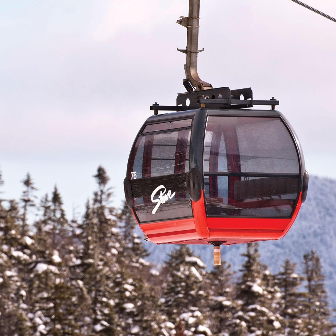 A red and black ski lift cabin numbered 76, flying above snowy pines, suggesting a ski getaway.