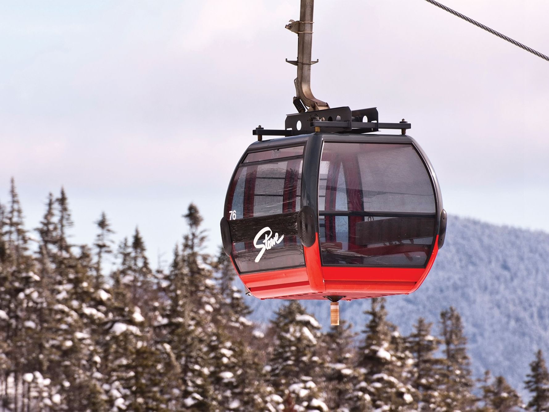 A red and black ski lift cabin numbered 76, flying above snowy pines, suggesting a ski getaway.