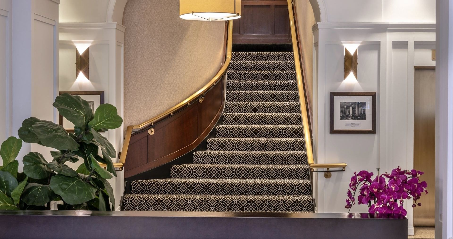 Reception lobby with a curved staircase, patterned carpet, and gold railings by a leafy plant at Warwick Allerton Chicago