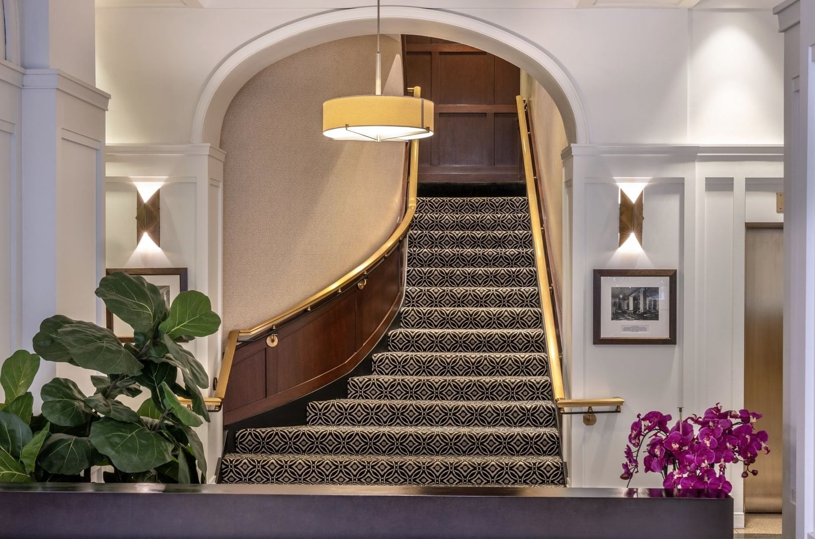 Reception lobby with a curved staircase, patterned carpet, and gold railings by a leafy plant at Warwick Allerton Chicago