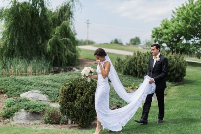 Groom holding the bride\'s flowing veil by garden shrubs, near a pond and trees at Mountain View Grand Resort & Spa