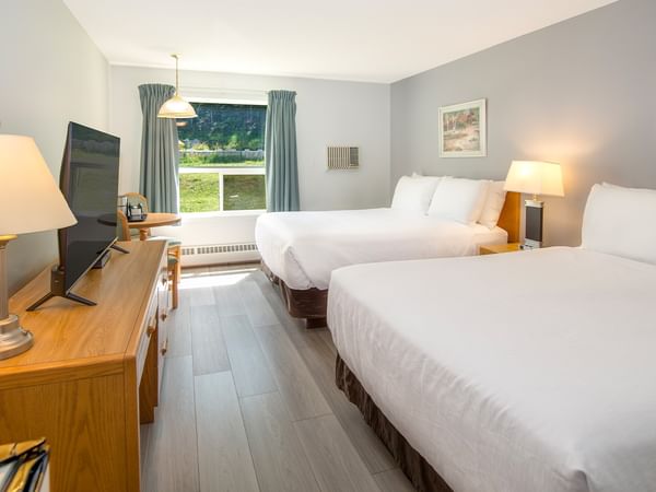 Two beds in a room with a TV, wooden dresser, and large window with curtains.