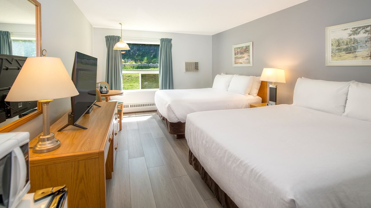 Two beds in a room with a TV, wooden dresser, and large window with curtains.