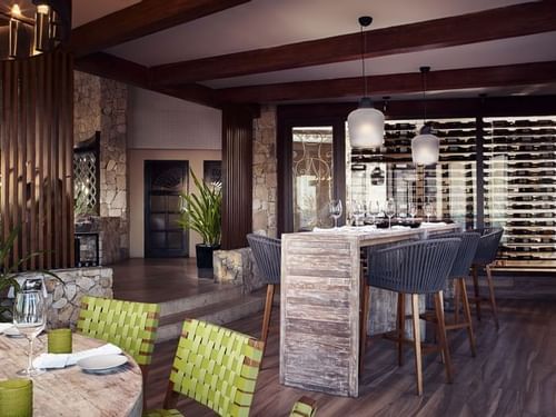 De Cortez Grill House dining area with a long wooden table, woven chairs, & a wine wall behind at Hacienda Del Mar Los Cabos