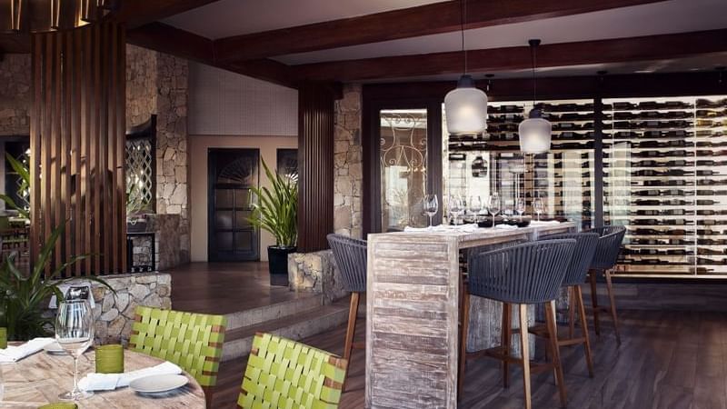 Comedor de De Cortez Grill House con una larga mesa de madera, sillas tejidas y una vinoteca detrás en Hacienda Del Mar Los Cabos