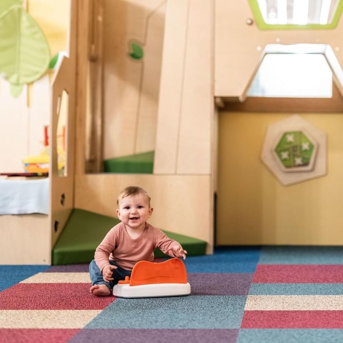 Baby spielt auf buntem Teppichboden mit Spielzeugauto in Spielbereich bei Falkensteiner Family Resort Sicily